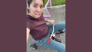 Adult riding a small red tricycle with a funny nervous expression, wearing casual clothes on a driveway near a toy soccer goal.